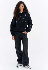 Černá mikina s kapucí s květinovou výšivkou v růžové a bílé barvě, vybavená přední kapsou, stahovací kapucí a volnějším střihem, kombinovaná s tmavými džínami.