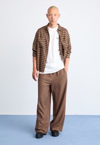 Brown check patterned shirt over a white t-shirt, paired with loose brown plaid pants. Black shoes and a plain background complete the look.