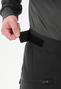 Svarta byxor med ett Velcro-justeringsband i midjan, med en ficka med dragkedja. Materialet verkar slätt och är utformat för funktionalitet.