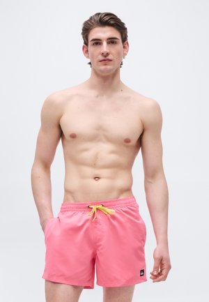 Jeune homme aux cheveux bruns courts portant un short de bain rose avec un cordon jaune, debout devant un fond clair uni.