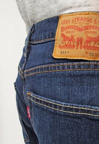 Sinisiä denim farkkuja, joissa on nahkainen lappu, jossa on oranssia tekstiä. Slim fit -malli, näkyvät saumat ja pieni punainen logo takataskussa.