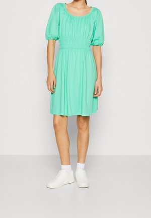 Femme portant une robe vert menthe longueur genou avec manches bouffantes, taille froncée, chaussettes blanches et baskets blanches, debout sur un sol gris.