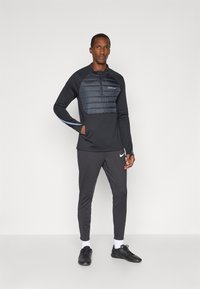 Must värvitud sportlik särk, millel on polsterdatud osa ja lukuga, koos tumeda halli pükstega. Omab valget Nike logo ja sujuvat tekstuuri.