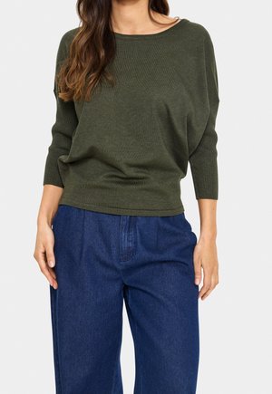Femme portant un pull en maille vert olive à manches longues avec des poignets côtelés et un pantalon en denim bleu ample, debout avec les mains détendues.