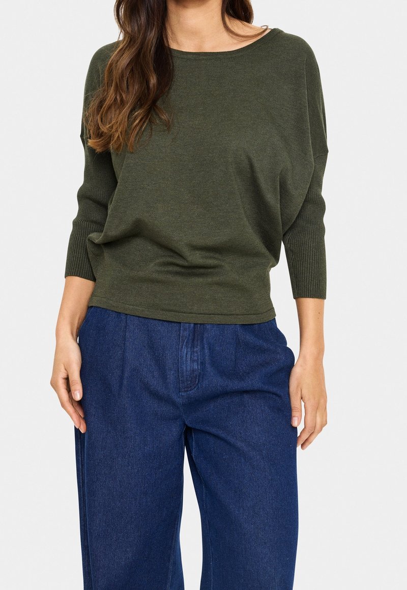 Femme portant un pull en maille vert olive à manches longues avec des poignets côtelés et un pantalon en denim bleu ample, debout avec les mains détendues.