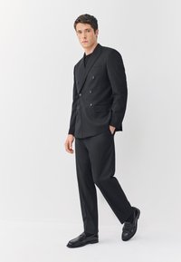 Fekete kétsoros zakó hegyes gallérral, hozzáillő fekete nadrág, valamint fényes fekete bőr cipő, fehér háttér előtt állva.