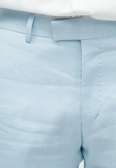 Κοντινή λήψη στη μέση μπλε λινών παντελονιών με θηλιές για ζώνη και κρυφό φερμουάρ, φορεμένες με ένα λευκό πουκάμισο που έχει μπει μέσα.