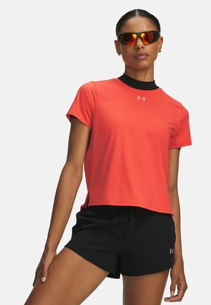 Frau mit verspiegelter Sportsonnenbrille, leuchtend korallfarbenem Sport-T-Shirt und schwarzen Shorts steht selbstbewusst vor schlichtem Hintergrund.