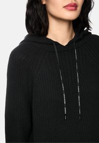 Schwarzer Hoodie mit strukturierter Oberfläche, ausgestattet mit einer vorderen Kängurutasche, Kordeln mit Logo-Akzenten und einem entspannten Design.