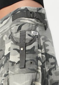 Grå kamuflasje cargo shorts med teksturert stoff, metallakcenter, justerbar midje og en synlig merkevarelapp. Har flere lommer.