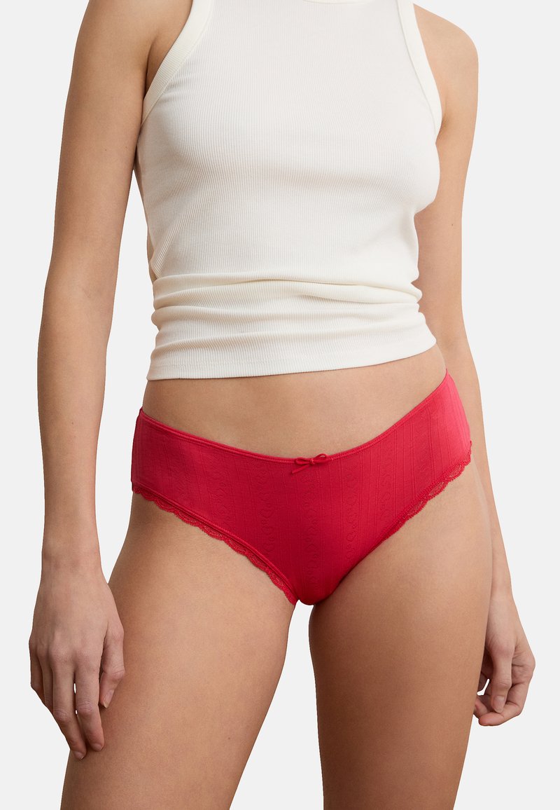 Frau trägt ein weißes ärmelloses Oberteil und rote Unterwäsche mit Spitzenbesatz, steht mit entspannten Armen seitlich am Körper vor weißem Hintergrund.