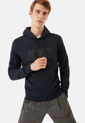 Young man wearing a black hoodie with drawstrings and gray plaid pants, standing with one hand near the hoodie collar.