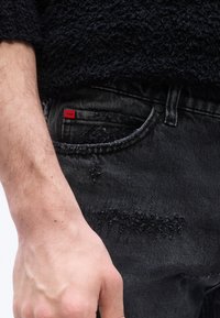 Αριστερό χέρι ακουμπισμένο δίπλα σε μαύρο τζιν με ξεθωριασμένο εφέ και κόκκινη ετικέτα "Hugo" στην μικρή μπροστινή τσέπη και μαύρο υφασμάτινο πουλόβερ από πάνω.