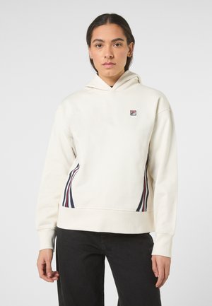 Jonge vrouw draagt een crème kleurige hoodie met een klein logo op de borst en strepen aan de zijkant, gecombineerd met een zwarte broek, en staat tegen een effen achtergrond.