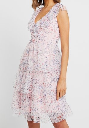 Woman wearing a sleeveless, knee-length, layered dress with pink and blue floral patterns on sheer white fabric.