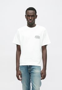 Valge puuvillane T-särk lühikeste varrukatega, millel on ümara kaelusega ja vasakul rinnal väike trükitud logo; kombineeritud hele siniste teksadega.