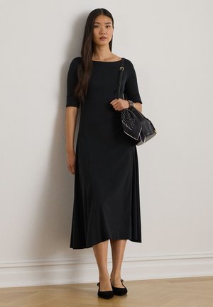 Vrouw draagt een zwarte midi-jurk met ellebooglange mouwen, houdt een zwarte handtas met een polkadot-sjaal vast, staat tegen een effen muur.