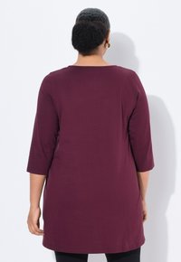 Burgunderfarbene Tunika mit Dreiviertelärmeln, aus weicher Stoff gefertigt, mit rundem Ausschnitt und längerer Silhouette.