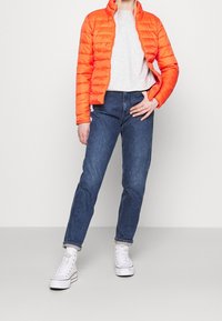Orangefarbene Steppjacke mit hohem Kragen, kombiniert mit einem grauen T-Shirt und blauen Jeans. Weiße High-Top-Sneaker runden das Outfit ab.