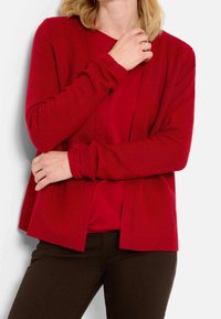 Cardigan rosso lavorato a maglia con apertura frontale, polsini a coste e tessuto testurizzato, indossato su una maglietta rossa coordinata, abbinato a pantaloni marroni.