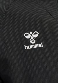 Un tessuto nero testurizzato presenta un logo bianco prominente con un design stilizzato di un'ape e la parola "hummel" qui sotto.