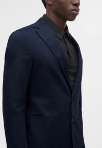 Homme portant un blazer bleu marine foncé par-dessus une chemise noire à col, visible de la poitrine au menton sur un fond clair uni.