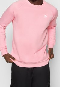 Růžová mikina s žebrovaným kulatým výstřihem, dlouhými rukávy a malým bílým logem Adidas na hrudi. Hladká textury látky.