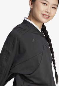 Črna športna majica s kapuco, ki ima tri črte na rami, visok ovratnik in subtilen Adidasov logotip. Mehak, teksturiran material.