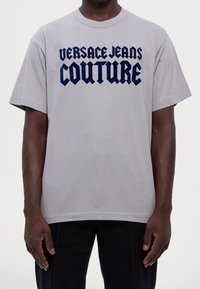 Uomo che indossa una maglietta grigia chiara con "VERSACE JEANS COUTURE" in grande font gotico blu scuro sul petto.