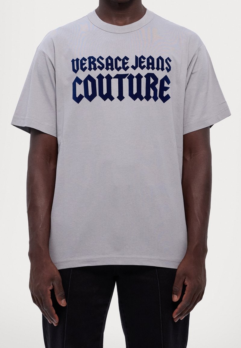 Uomo che indossa una maglietta grigia chiara con "VERSACE JEANS COUTURE" in grande font gotico blu scuro sul petto.