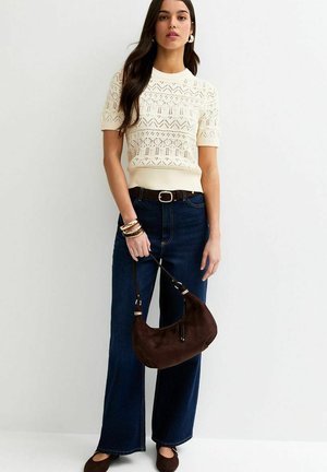 Vrouw draagt een crèmekleurige korte mouw schipperstrui, donkere wijde jeans met riem, houdt een bruine handtas vast en draagt armbanden en oorbellen.