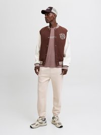 Bruine en witte varsityjack met gestreepte manchetten, beige sweatpants en een bruine pet, met een grafisch logo op de borst.