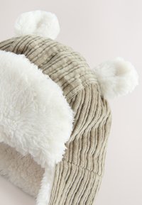 Béžová čiapka z kordurovej tkaniny s bielym umelým kožušinovým lemom, s zaoblenými úchytmi na ušiach a textúrovaným dizajnom. Navrhnutá pre teplo a pohodlie.