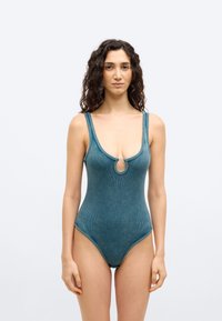 Γυναί κα που στέκεται και κοιτάζει μπροστά, φορώντας ένα μονόχρωμο ολόσωμο μαγιό σε τυρκουάζ χρώμα με ριμπ ύφασμα, κούμπωμα μπροστά και λεπτομέρεια με άνοιγμα.