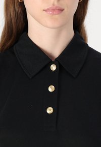 Svart ribbet polo skjorte med krage, med fire gullknapper foran, som fremhever det teksturerte stoffet og den strukturerte designen.