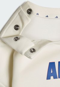 Detailný záber na krémovú látku s tromi kovovými cvočkami a čiastočným modrým a červeným nápisom na odeve.