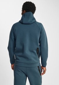 Felpa teal con colletto alto, polsini a coste e tasche laterali con zip. Tessuto morbido e testurizzato con vestibilità rilassata e schiena allungata.