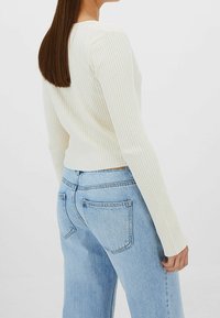 Hladký krémový top s dlouhým rukávem a přiléhavým siluetu, kombinovaný se světlými modrými džínami s vysokým pasem. Viditelné švy a rovný střih nohavic.