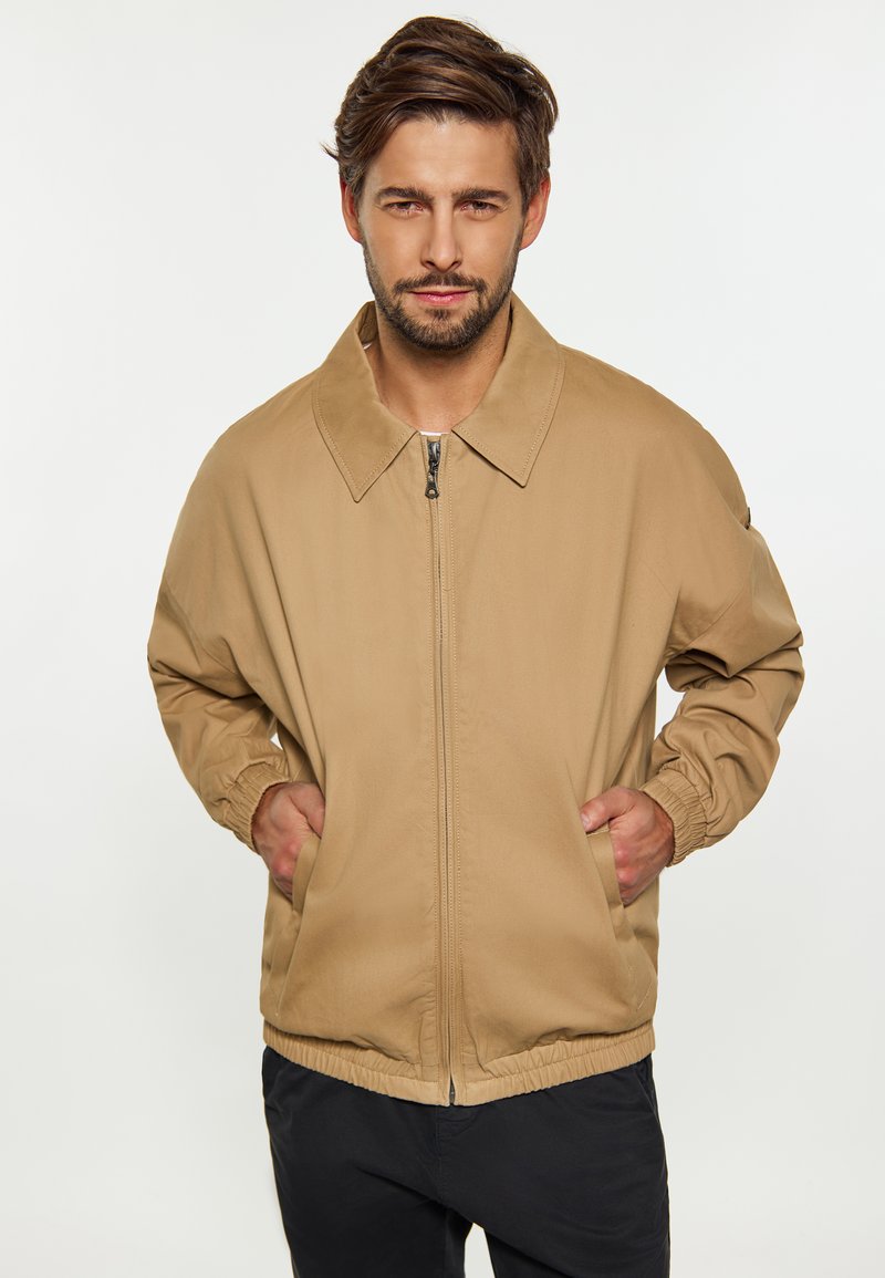 Beige Nylonjacke mit einem Reißverschluss vorne, großem Kragen, elastischen Bündchen und Taille. Verfügt über zwei seitliche Taschen und ein lässiges Design.