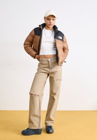 Hnědá a černá bunda typu puffer, bílý crop top, světle béžové roztrhané džíny a tmavě modré boty. Bílá čepice a stříbrné šperky. Žluté pozadí.