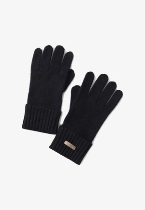 Grüner Pullover, schwarze Strickmütze und Schal, schwarze Handschuhe, beige Hose und weiße Sneakers. Der Pullover hat gerippte Bündchen und Struktur.