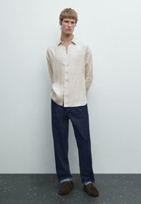 Linen shirt in beige with a button-up front, paired with dark blue denim pants. The model wears brown shoes, standing against a neutral background.
