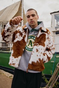 Mladý muž v hnědé a bílé bundě z umělé kožešiny a šedé mikině s logem Oakland Athletics stojí poblíž zelené a bílé lodi s plachtami.