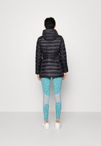 Svart vadderad jacka med huva och horisontella sömmar. Blå leggings med ormmönster och genomskinliga partier. Vita sneakers med ren design.