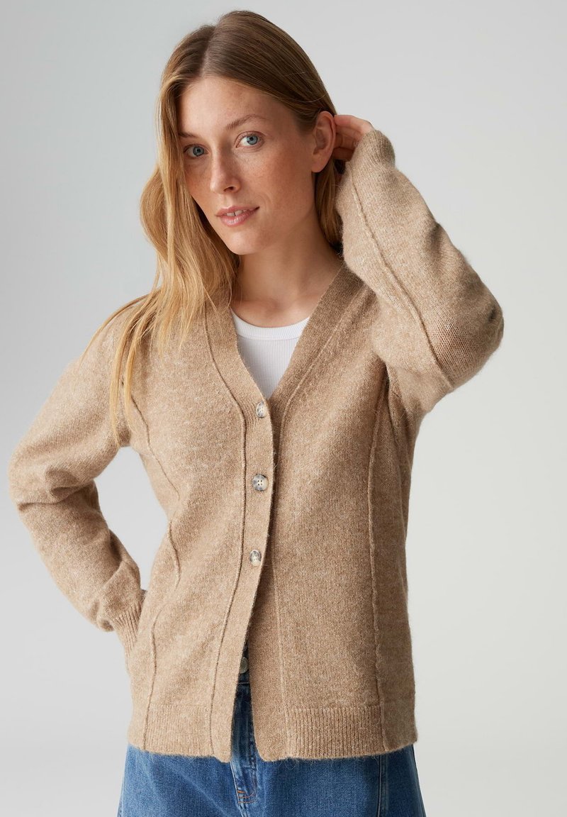 Cardigan in lana beige con cinque bottoni, scollo a V, polsini a coste e cuciture laterali. Indossato sopra una maglietta bianca e jeans blu.