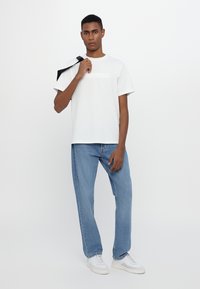 White cotton t-shirt with vertical branding on the chest, paired with light blue denim jeans and white sneakers. Model holds a bag.