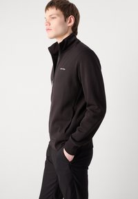 Melns zip-up džemperis ar augstu apkakli, gluda tekstūra un Calvin Klein logo kreisajā krūtī. Modelis to valkā, rokas kabatās.