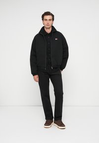 Black padded bomber jacket with a high collar, zip closure, and logo patch, paired with black trousers and brown sneakers.