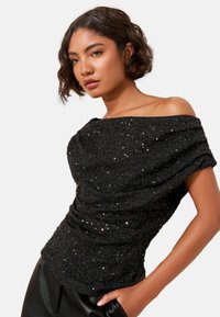Čierny off-the-shoulder top s textúrovanými flitrami, drapovanou látkou a priliehavým strihom, v kombinácii s elegantnými čiernymi nohavicami.