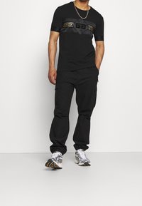 Schwarzes T-Shirt mit einem glänzenden Schwarz- und Goldmuster auf der Brust, kombiniert mit lockeren schwarzen Cargo-Hosen und grauen Sneakers. Silbergeldetail.